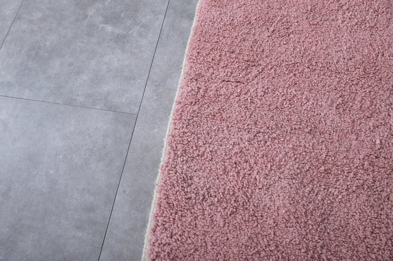 May include: Close-up of a soft, pink, fluffy rug with a white border. The rug is placed on a gray tiled floor.