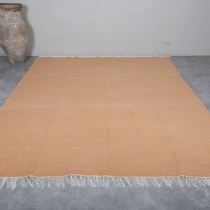 May include: A large, rectangular, peach-colored rug with white fringe along the edges. The rug is lying flat on a gray tiled floor.