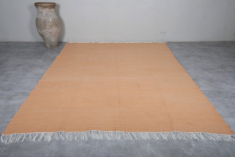 May include: A large, rectangular, peach-colored rug with white fringe along the edges. The rug is lying flat on a gray tiled floor.