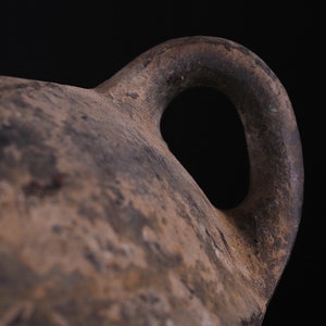 May include: Close-up of a brown clay pot with a single handle. The handle is thick and rounded, and the pot has a rough, textured surface.