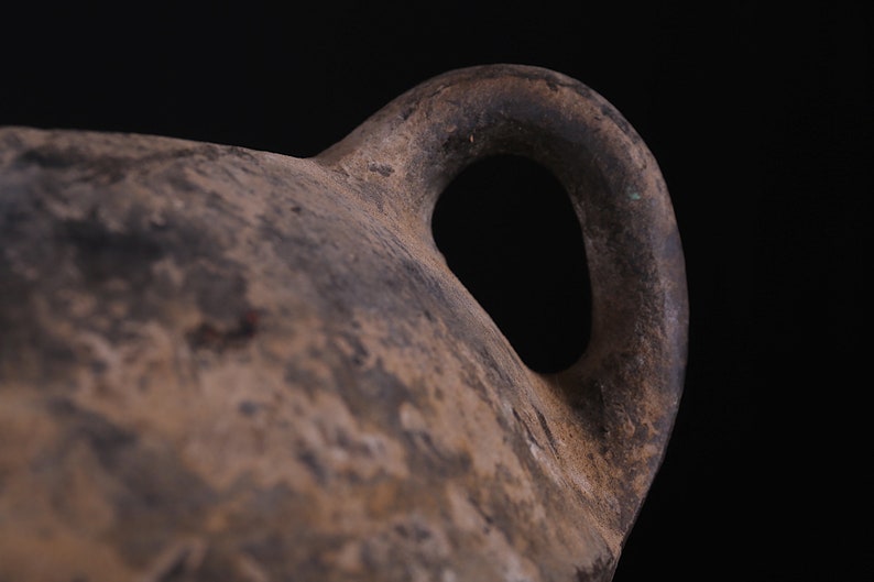 May include: Close-up of a brown clay pot with a single handle. The handle is thick and rounded, and the pot has a rough, textured surface.