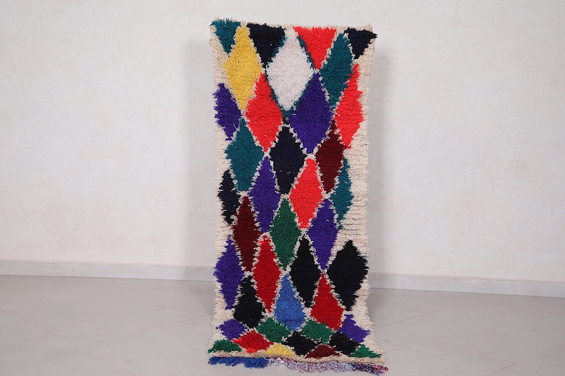 May include: A colorful Moroccan rug with a geometric diamond pattern in shades of red, blue, green, yellow, and white. The rug has a fringe border and is made of wool.
