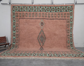 Puede incluir: Una alfombra grande y rectangular con fondo rosa y un patrón geométrico verde y negro. La alfombra tiene un borde de flecos.