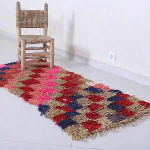May include: A long, rectangular rug with a checkered pattern in shades of pink, red, brown, and blue. The rug has a fringe border and is made of a soft, fluffy material. A wooden chair sits on the rug.