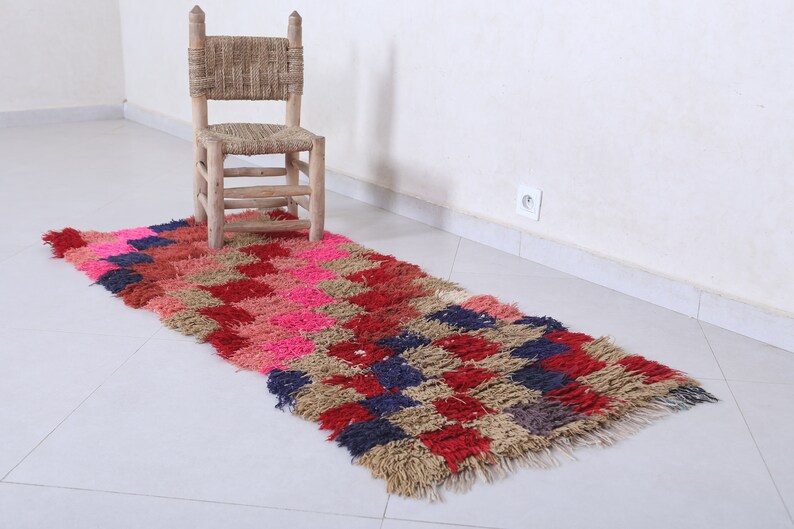 May include: A long, rectangular rug with a checkered pattern in shades of pink, red, brown, and blue. The rug has a fringe border and is made of a soft, fluffy material. A wooden chair sits on the rug.