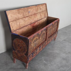 May include: An antique wooden chest with an open lid, featuring detailed carvings and a distressed finish. The chest is painted in red, brown, and blue, with decorative circular patterns. It stands on four short legs, offering storage.