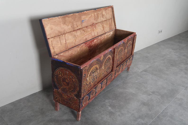 May include: An antique wooden chest with an open lid, featuring detailed carvings and a distressed finish. The chest is painted in red, brown, and blue, with decorative circular patterns. It stands on four short legs, offering storage.
