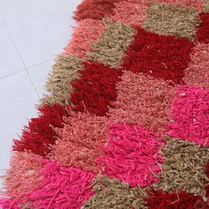 May include: A colourful rug with a geometric pattern in shades of pink, red, and brown. The rug is made of fluffy, textured yarn and has a diamond-shaped design.