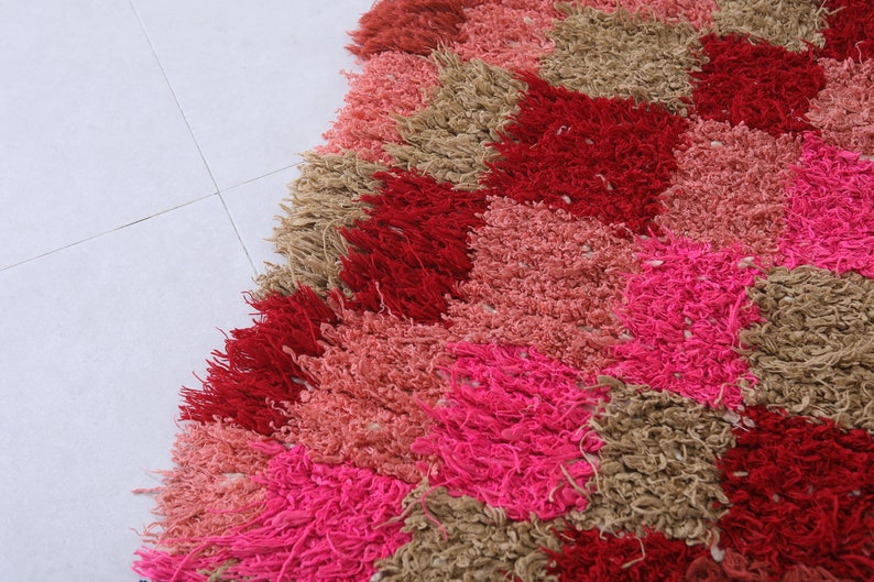 May include: A colourful rug with a geometric pattern in shades of pink, red, and brown. The rug is made of fluffy, textured yarn and has a diamond-shaped design.