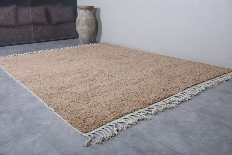 May include: A light brown shag rug with a white fringe border. The rug is laid out on a gray tiled floor.