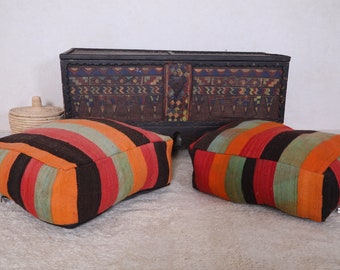 May include: Two colorful striped floor cushions with a dark wooden chest with geometric patterns in the background.