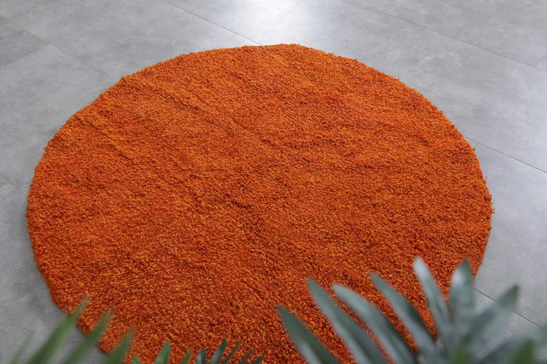 May include: A round, orange-colored rug with a fluffy texture. The rug is made of a soft, plush material and has a subtle, circular pattern.