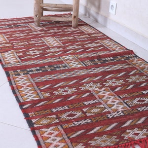 May include: A red, black, and white patterned rug with fringe. The rug has a geometric design and is made of wool.