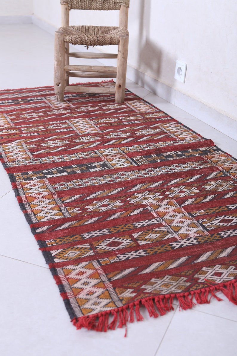 May include: A red, black, and white patterned rug with fringe. The rug has a geometric design and is made of wool.