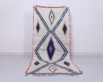 May include: A white Moroccan rug with a geometric pattern in shades of blue, red, and yellow. The rug has a fringe border.