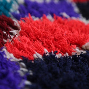May include: Close-up of a colorful, textured rug with red, blue, white, and green hues. The rug is made of fluffy, woven fibers.