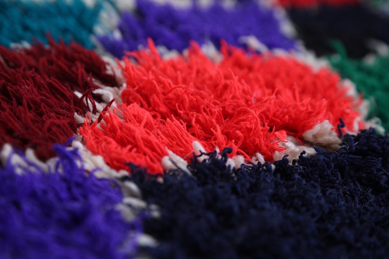 May include: Close-up of a colorful, textured rug with red, blue, white, and green hues. The rug is made of fluffy, woven fibers.