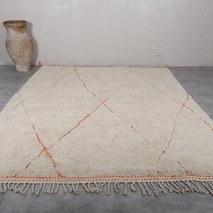 May include: Cream-colored, rectangular rug with a geometric design. The rug features orange diamond patterns and a fringed edge. A large, tan vase is in the background.