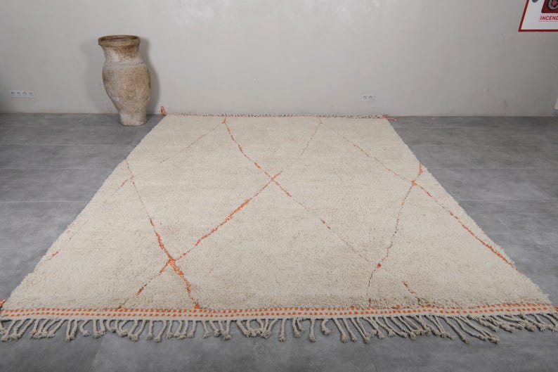 May include: Cream-colored, rectangular rug with a geometric design. The rug features orange diamond patterns and a fringed edge. A large, tan vase is in the background.
