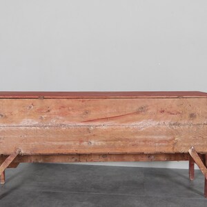 May include: A rustic wooden chest with a red-painted top and legs. The chest is made of weathered wood, showing visible grain and knots. The legs are angled for support. A vintage storage solution.
