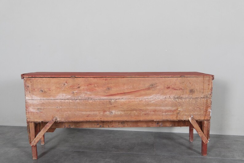 May include: A rustic wooden chest with a red-painted top and legs. The chest is made of weathered wood, showing visible grain and knots. The legs are angled for support. A vintage storage solution.