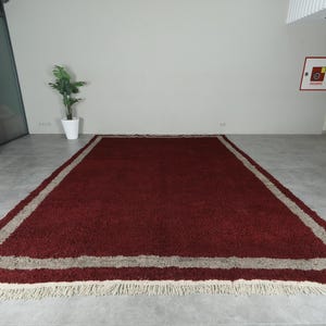 May include: A rectangular, burgundy-colored rug with a beige border and fringed edges. The rug is placed on a gray floor, and the room has white walls. The rug's dimensions are not visible.