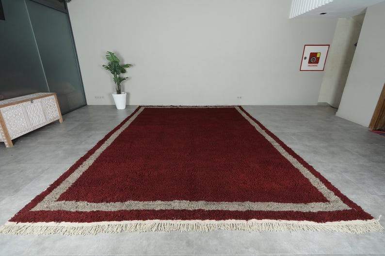 May include: A rectangular, burgundy-colored rug with a beige border and fringed edges. The rug is placed on a gray floor, and the room has white walls. The rug's dimensions are not visible.