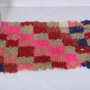 May include: A colourful, rectangular rug with a checkered pattern in shades of pink, red, brown, and blue. The rug is made of a fluffy, textured material.
