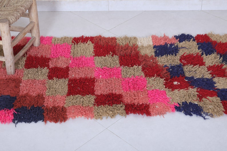 May include: A colourful, rectangular rug with a checkered pattern in shades of pink, red, brown, and blue. The rug is made of a fluffy, textured material.