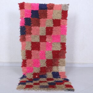 May include: A long rectangular rug with a checkered pattern in shades of pink, red, beige, and blue. The rug is made of a soft, fluffy material.