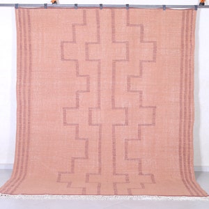 May include: A light pink rug with a geometric pattern in a darker shade of pink. The rug has a fringe border.