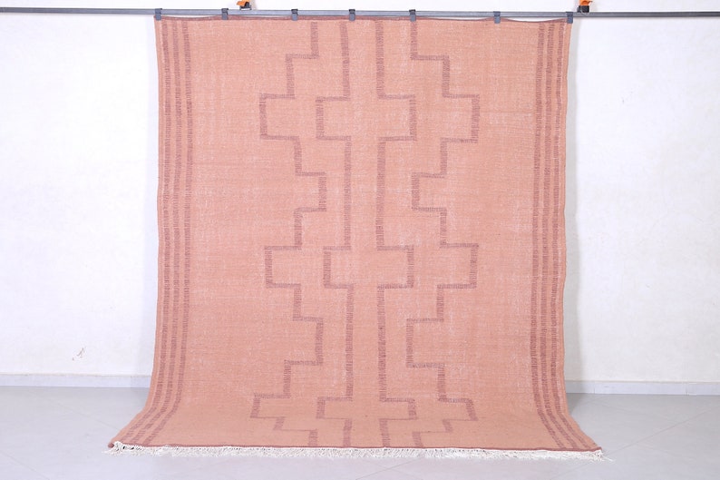 May include: A light pink rug with a geometric pattern in a darker shade of pink. The rug has a fringe border.