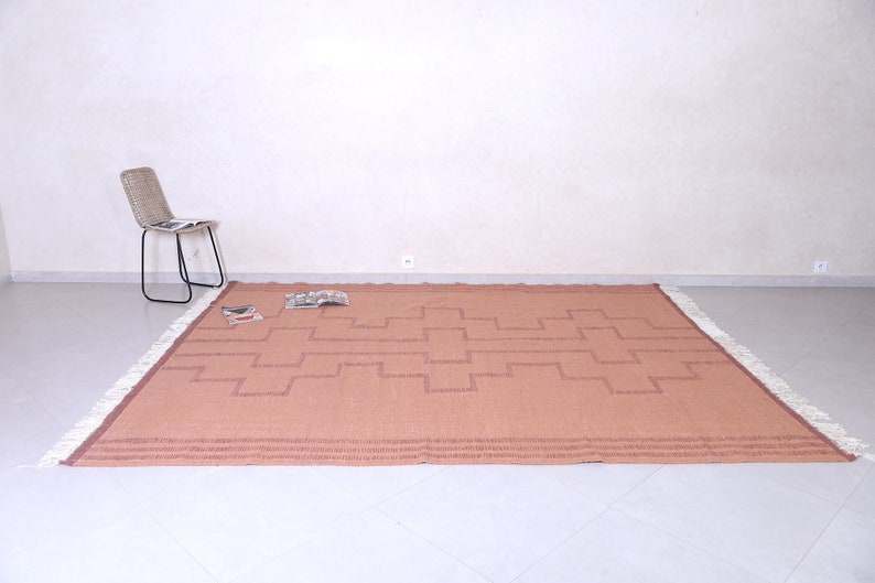 May include: A light brown woven rug with a geometric pattern and white fringe. The rug is laid out on a white tiled floor.