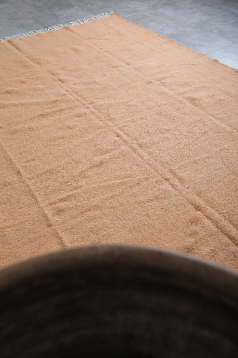 May include: A light brown rug with a textured surface. The rug has a fringe border and is laid out flat on a gray floor.