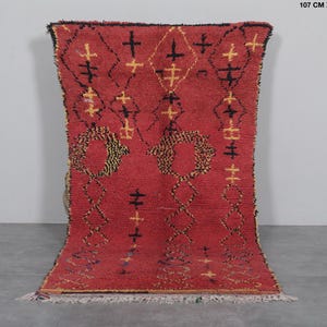 May include: A red Moroccan rug with a geometric pattern of black and yellow crosses and circles. The rug is 3.5 feet by 7 feet and is made of wool.