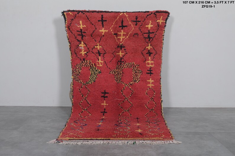 May include: A red Moroccan rug with a geometric pattern of black and yellow crosses and circles. The rug is 3.5 feet by 7 feet and is made of wool.