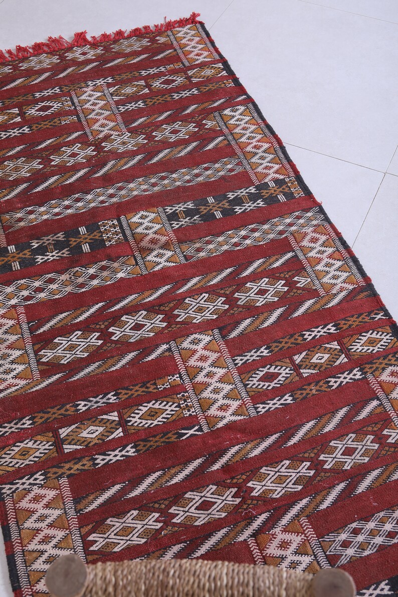 May include: A red, brown, and white patterned rug with geometric designs. The rug has a fringe on the top edge.