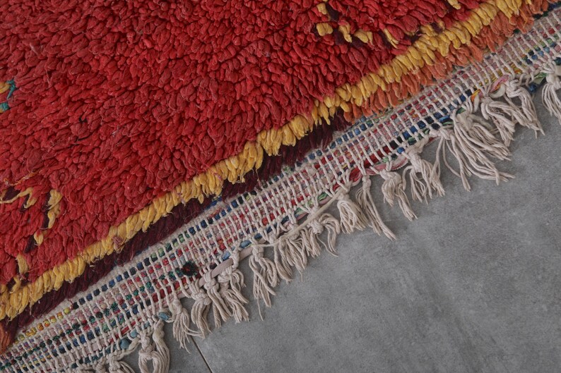 May include: A red, yellow, and white Moroccan rug with a fringe border. The rug has a textured, fluffy surface.