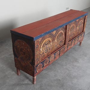 May include: A long, rectangular wooden chest with a red top and intricate painted designs. The chest features ornate patterns in gold, blue, and brown, with decorative elements on the front and sides. It stands on four short legs.