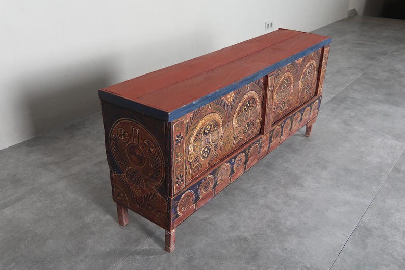 May include: A long, rectangular wooden chest with a red top and intricate painted designs. The chest features ornate patterns in gold, blue, and brown, with decorative elements on the front and sides. It stands on four short legs.
