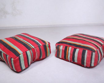 May include: Two red, green, black, and white striped floor cushions. The cushions are made of woven fabric and have a square shape.