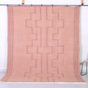 May include: A blush pink rug with a geometric pattern woven into the fabric. The rug has a fringe border.