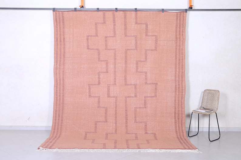 May include: A blush pink rug with a geometric pattern woven into the fabric. The rug has a fringe border.