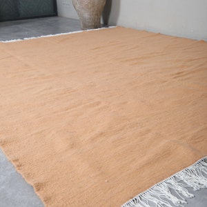 May include: A large, rectangular, light orange rug with white fringe along the edges. The rug is lying flat on a gray tiled floor.