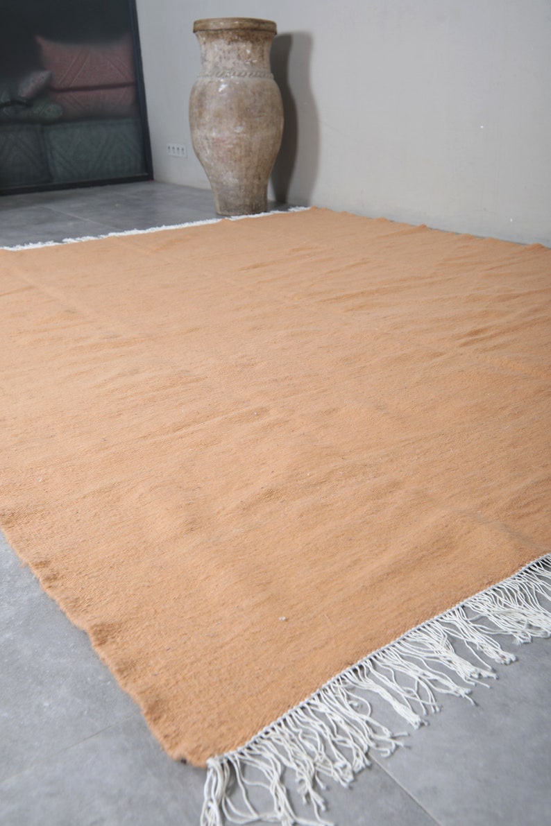 May include: A large, rectangular, light orange rug with white fringe along the edges. The rug is lying flat on a gray tiled floor.