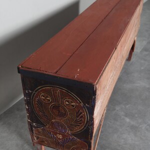May include: A long, rectangular wooden chest with a red-brown top and sides. The front of the chest features a circular, ornate design in shades of brown, gold, and blue. The chest rests on four short, red-brown legs.
