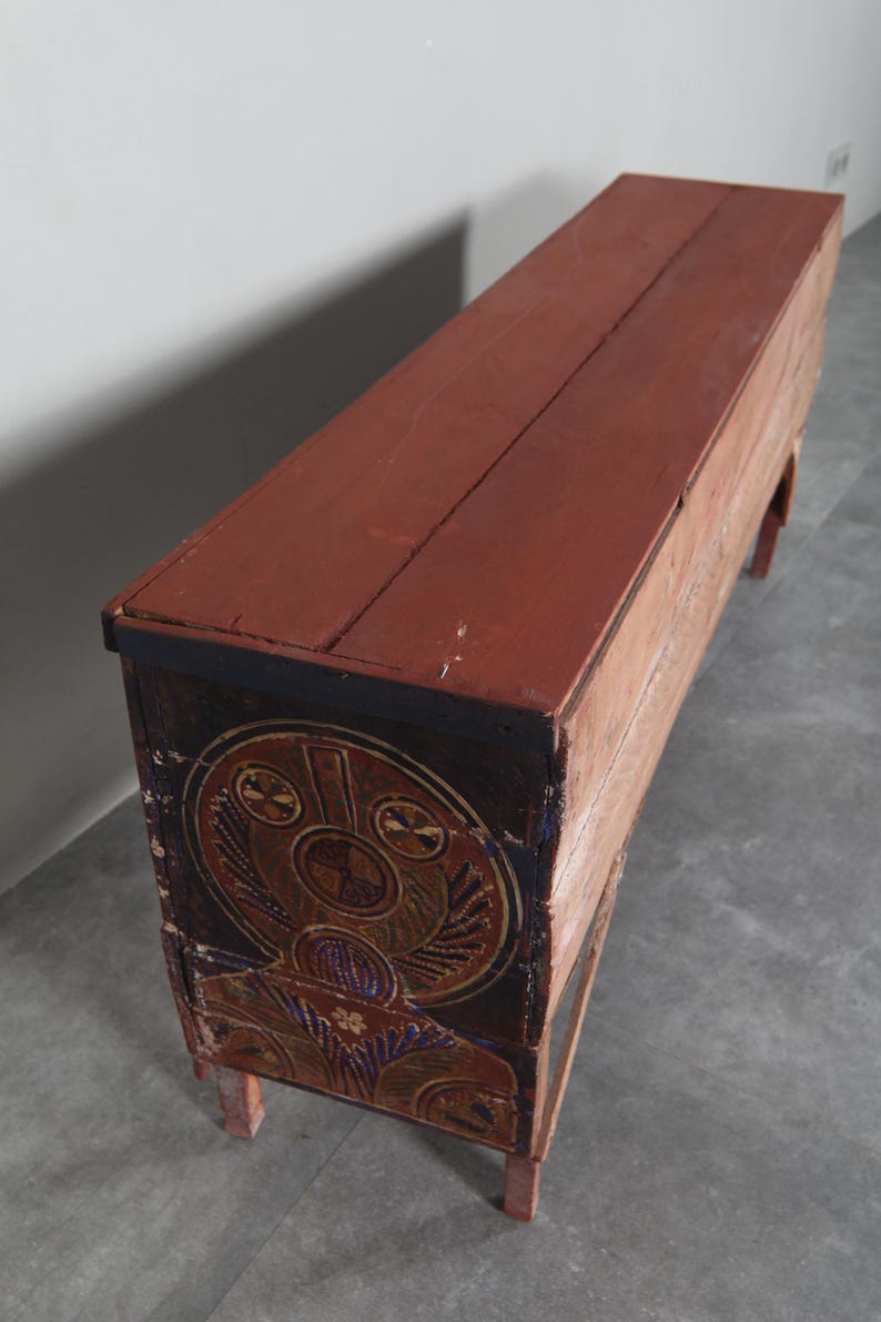 May include: A long, rectangular wooden chest with a red-brown top and sides. The front of the chest features a circular, ornate design in shades of brown, gold, and blue. The chest rests on four short, red-brown legs.