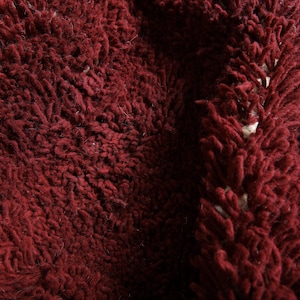May include: Close-up of a deep burgundy, shaggy textile. The fabric has a thick, textured pile with a dense, looped appearance. The color is a rich, dark red with subtle variations in tone, suggesting depth and dimension.