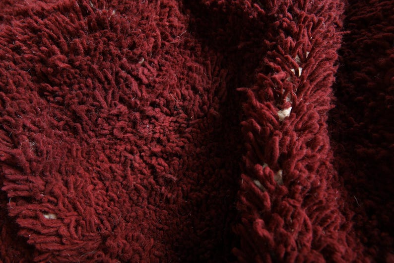 May include: Close-up of a deep burgundy, shaggy textile. The fabric has a thick, textured pile with a dense, looped appearance. The color is a rich, dark red with subtle variations in tone, suggesting depth and dimension.