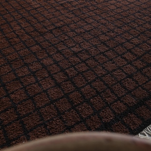 Peut inclure: Un tapis rectangulaire marron avec un motif quadrill&eacute; de lignes noires. Le tapis a une surface textur&eacute;e et moelleuse et une frange sur un bord. Le motif est un motif r&eacute;p&eacute;titif de carr&eacute;s.